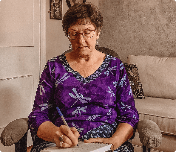 An elderly woman in a purple floral blouse writing in a notebook.